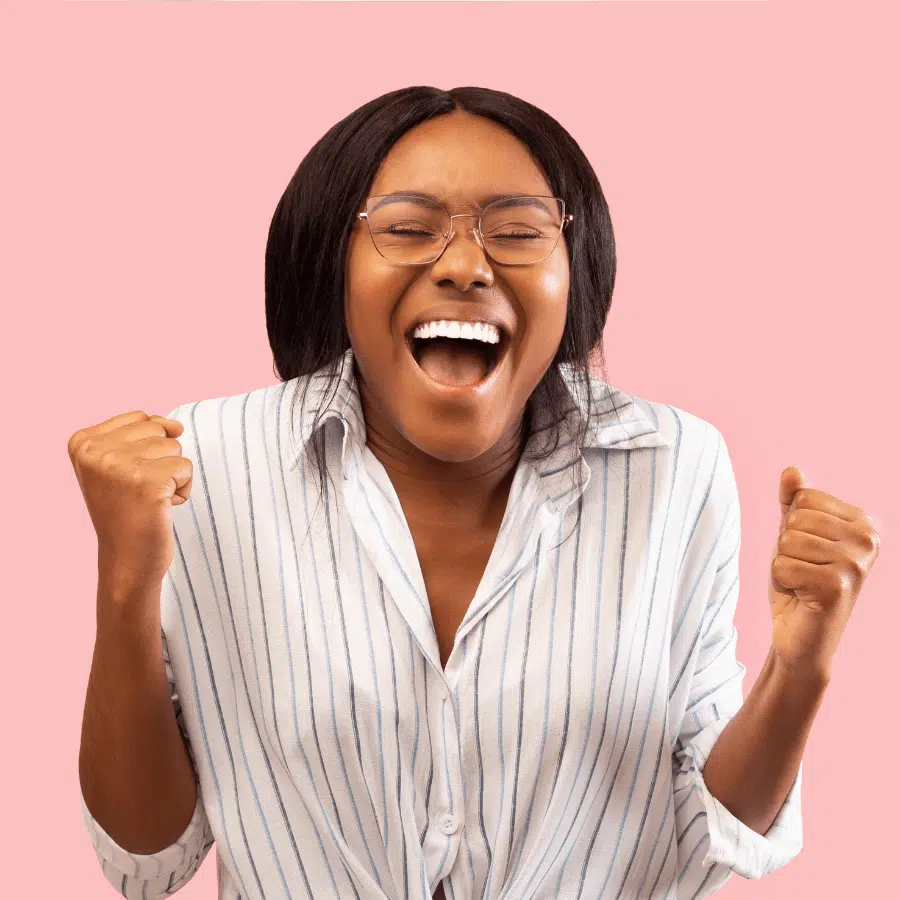 Happy young woman celebrating success with a big smile, wearing glasses and a striped shirt, against a pink background.