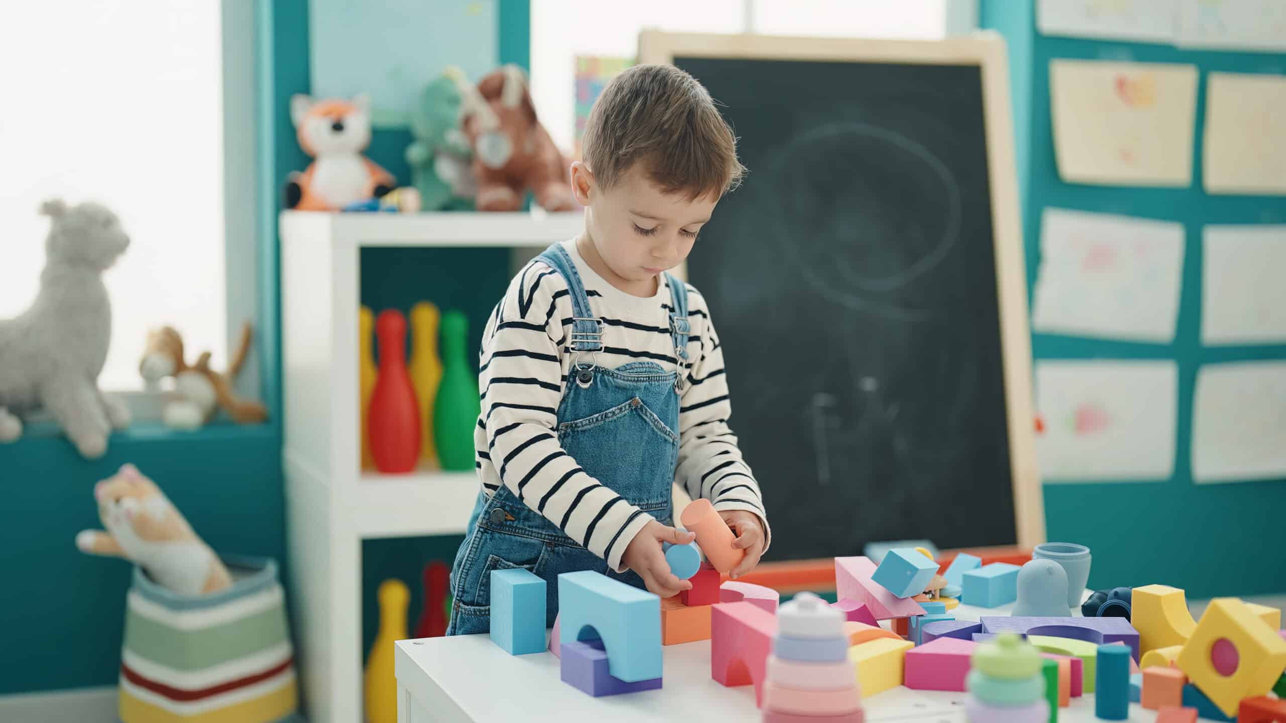 Toddler playing with colorful blocks in a playroom setting with toys and a chalkboard in the background.